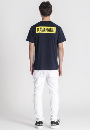 Mand iført en marineblå t-shirt med "KAVANAGH" i gul på ryggen, hvide bukser og sorte sneakers med hvide såler og branding.