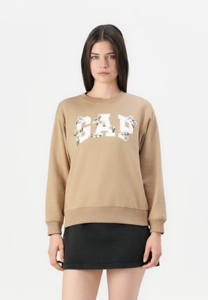 Nais, kes kannab beezhhi sweatshirt'i, millel on valged lilledega kaunistatud "GAP" kiri, musta seeliku kõrvale, seismas valge tausta ees.
