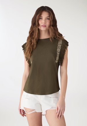 Donna con capelli castani ondulati che indossa un top verde oliva senza maniche con frange e rifiniture decorate sulle spalle, abbinato a pantaloncini bianchi strappati.