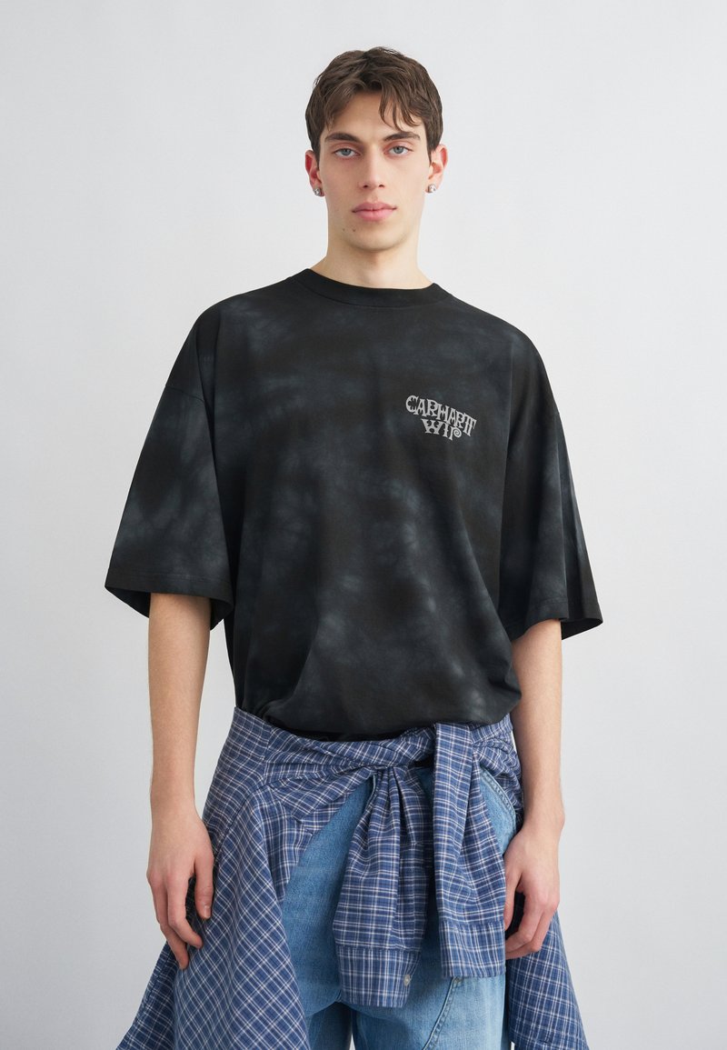 Νέος άνδρας με κοντά μαλλιά, φοράει μαύρο t-shirt Carhartt WIP με tie-dye και μπλε πουκάμισο με καρό που έχει δέσει γύρω από τη μέση του, μπροστά σε απλό φόντο.