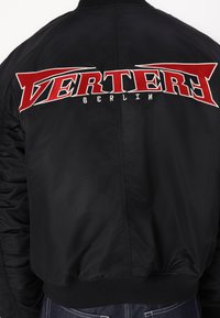 Črna bomberska jakna z velikim rdečim in belim vezenim logotipom "VERTER" in "BERLIN" na hrbtni strani, z rebrastimi manšetami in robom.