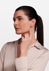 Strieborný prsteň s dizajnom kvetu, ktorý má päť okvetných lístkov, doplnený centrálnym kameňom. Modelka pózuje s rukou blízko tváre a nosí béžovú blúzku.
