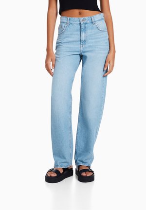 Femme portant un jean droit bleu clair, un crop top noir, et des sandales à plateforme noires avec des boucles, debout devant un fond blanc.
