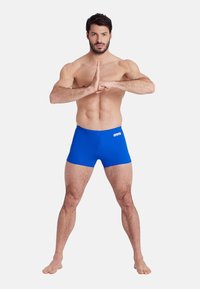 Bañador azul hecho de material elástico, diseño ajustado, con un logo blanco en la parte frontal y una textura suave. El modelo posa con las manos juntas.
