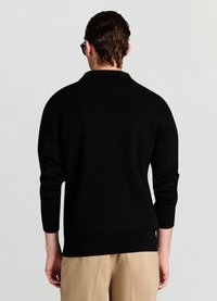 Pull noir avec un col classique, des poignets et un ourlet côtelés. La vue de dos révèle une texture lisse et une coupe ajustée, associée à un pantalon beige clair.