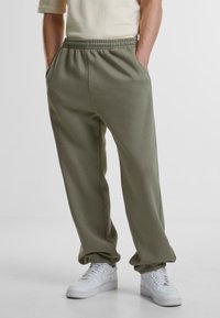 Olivenfargede sweatpants med elastisk midje, sidelommer og elastiske mansjetter, sammen med hvite joggesko. Myk, glatt tekstur.