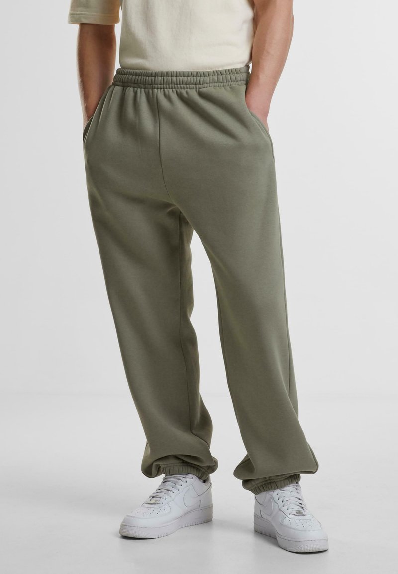 Olivenfargede sweatpants med elastisk midje, sidelommer og elastiske mansjetter, sammen med hvite joggesko. Myk, glatt tekstur.