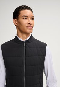 Junger Mann mit kurzem lockigem Haar, der eine schwarze gesteppte Steppweste über einem weißen Langarmshirt trägt und leicht nach rechts blickt, vor einem neutralen Hintergrund.