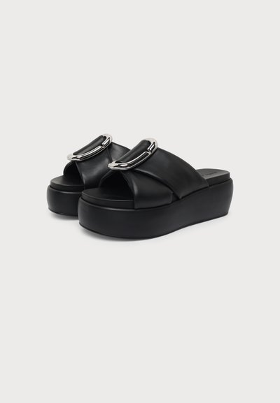 Paire de sandales à plateforme noires avec brides matelassées et détails de boucles argentées brillantes sur un fond clair.