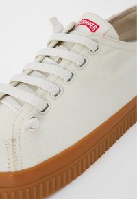 Beige canvas sneaker with beige laces, featuring a red logo tag and a textured brown rubber sole. Simple design with metal eyelets.