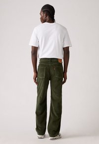 Man met gevlochten haar staat met zijn rug naar de camera gekeerd in een wit T-shirt, olijfgroene corduroy broek en witte sneakers tegen een effen achtergrond.