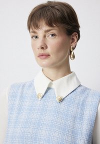 Haine din tweed albastru cu guler alb, având două butoane decorative aurii. Materialul are un model texturat. Cercei aurii în formă de lacrimă.