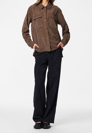 Femme portant une chemise en velours côtelé marron à boutons avec poches poitrine, pantalon large en velours côtelé noir et chaussures noires, debout les mains dans les poches.
