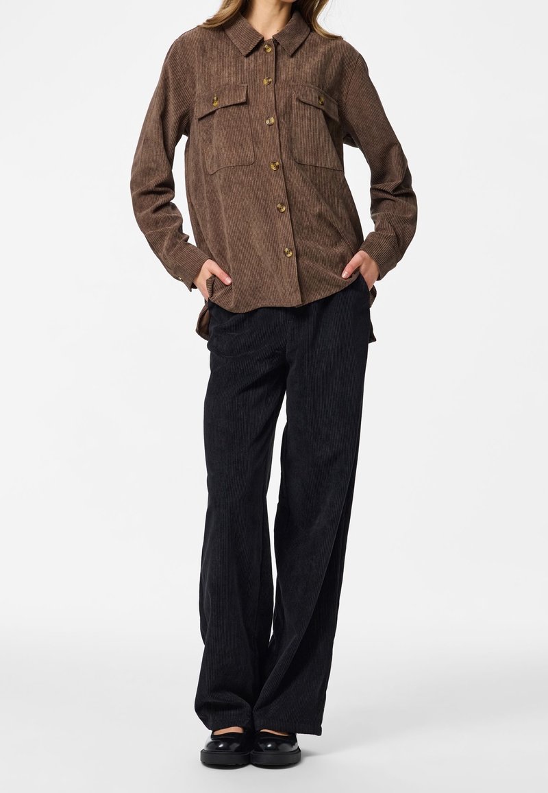 Femme portant une chemise en velours côtelé marron à boutons avec poches poitrine, pantalon large en velours côtelé noir et chaussures noires, debout les mains dans les poches.