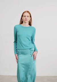 Haut à manches longues teal associé à une jupe satin teal avec des détails froncés sur les côtés. Texture lisse, design ajusté et style minimaliste.