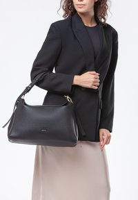 Borsa a tracolla nera in pelle con superficie strutturata, dettagli in metallo dorato, tracolla removibile e forma arrotondata, tenuta da una persona in giacca nera.
