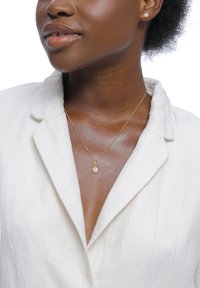 Goldene Halskette mit kleinem rundem Anhänger, der mit einem blassen Stein besetzt ist, getragen über einer strukturierten weißen Bluse mit Kragen.