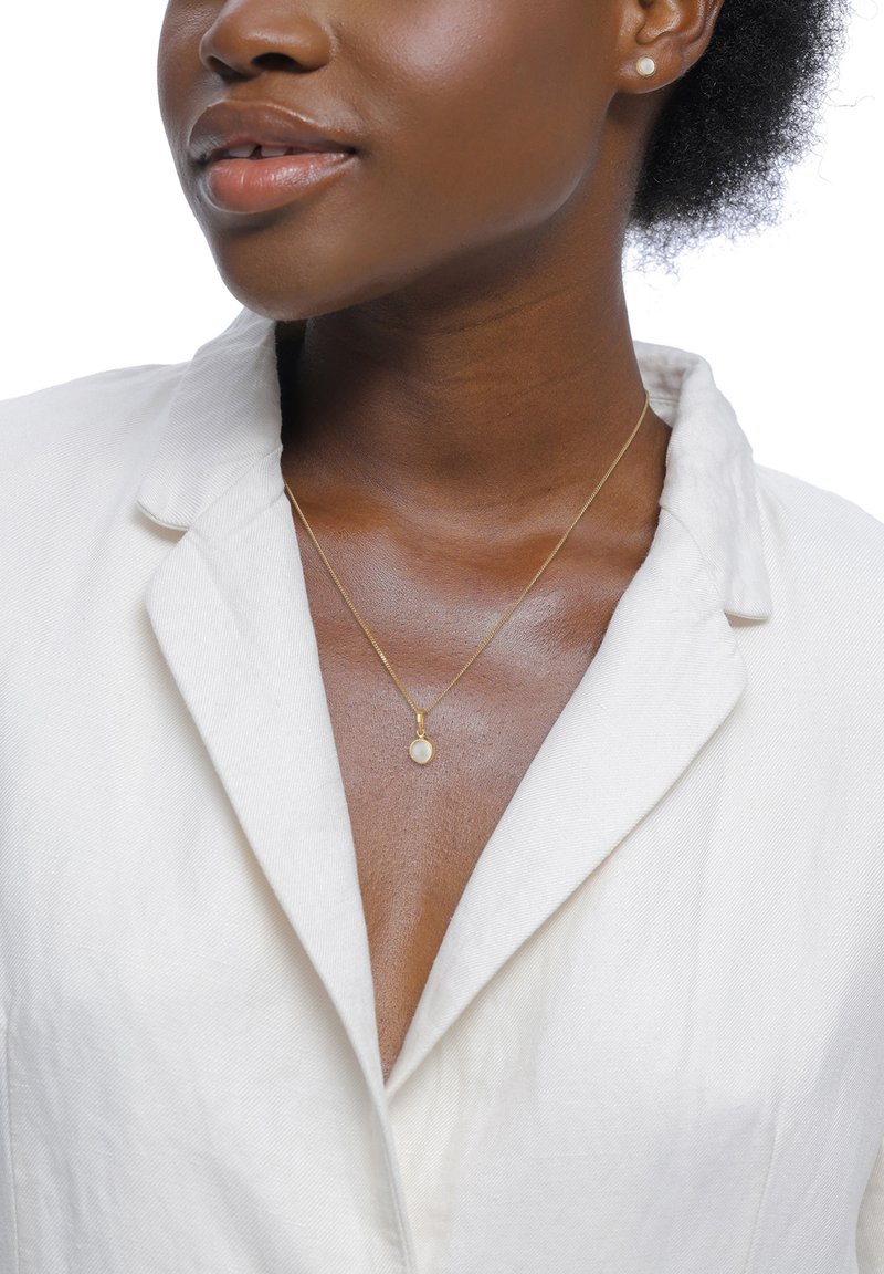 Goldene Halskette mit kleinem rundem Anhänger, der mit einem blassen Stein besetzt ist, getragen über einer strukturierten weißen Bluse mit Kragen.