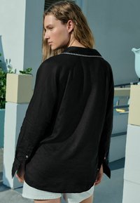 Cămașă neagră din in, cu mâneci lungi, manșetă cu nasture și o cusătură decorativă pe guler. Prezentată cu pantaloni scurți deschiși.