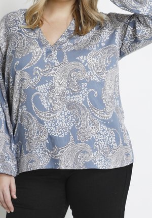Femme portant un chemisier bleu à manches longues avec motif cachemire et un pantalon noir, la main levée près de la tête.