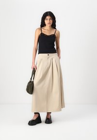 Svart topp med blonder, sammen med en beige, plisserte ankellange skjørt. Modellen holder en grønn veske og har på seg svarte platåsko.