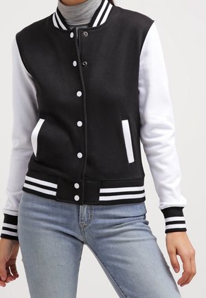 Veste universitaire noire et blanche avec col, poignets et ourlet rayés, portée par-dessus un col roulé gris et un jean bleu clair.