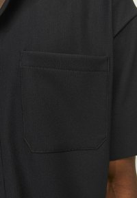 Black short-sleeve shirt made of ribbed fabric, featuring a left chest pocket and subtle stitching detail.