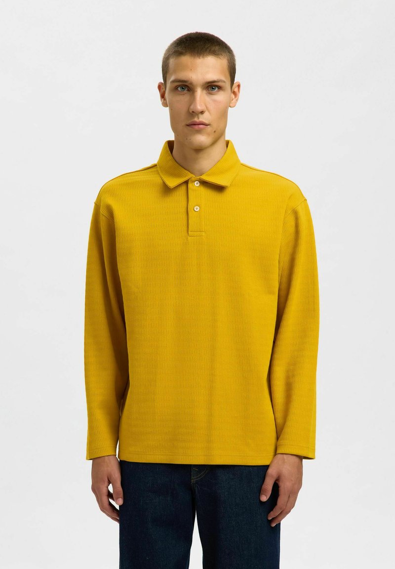 Langarm-Polohemd in leuchtendem Gelb, mit strukturiertem Muster, Drei-Knopf-Leiste und klassischem Kragen. Kombiniert mit dunklen Jeans.