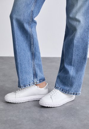 Białe niskie sneakersy z teksturowaną podeszwą noszone z jasnoniebieskimi prostymi dżinsami na szarej podłodze.