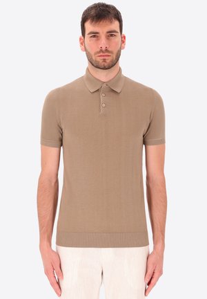 Uomo con capelli corti e scuri che indossa una maglietta polo beige a maniche corte e pantaloni chiari, in piedi davanti a uno sfondo uniforme.