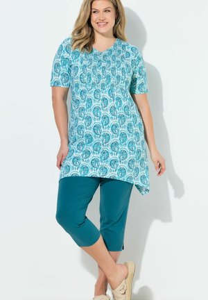 Uśmiechnięta kobieta w niebieskiej tunice w wzory z krótkimi rękawami, połączona z zielonkawymi, skróconymi spodniami i beżowymi butami wsuwanymi, stoi na białym tle.