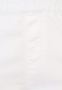 Bílá džínovina s hladkým povrchem, zdobená tónováním švů a detaily přední kapsy. Viditelný vzor tkaní.