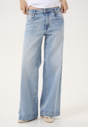 Hellblaue Jeans mit weitem Bein, getragen mit weißen Schuhen und einem weißen Oberteil, Person hält den Bund leicht nach unten gezogen.