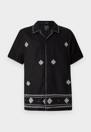 Svart kortermet skjorte laget av stoff med hvite broderte mønstre og detaljer. Har en knapp-oppe foran og dekorativ søm.