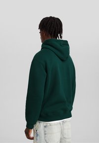 Zelená mikina s kapucí na stažení, žebrovanými manžetami a lemem. Zadní část ukazuje hladkou texturu a pohodlný střih, kombinovaná s světlými džínami.