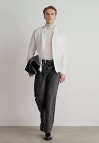 Model trägt ein weißes Hemd über einem beigen Rollkragenpullover, kombiniert mit lockeren, dunklen Jeans und schwarzen Schuhen, während sie eine schwarze Tasche hält.