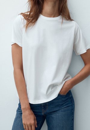 Vrouw draagt een effen witte T-shirt met korte mouwen en kantdetails op de schouders, gecombineerd met een blauwe spijkerbroek, staand tegen een lichte muur.