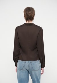 Inimene kannab tumedat pruuni sobiva lõikega pikkade varrukatega pluusi, mille mansetid on nööbiga kinni, ja helesiniseid kõrge vöökohaga teksapükse, seistes valgel taustal seljaga.