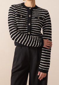Randig svart och vit cardigan med strukturerad stickning, rundade knappar, långa ärmar och kort längd, tillsammans med svarta byxor.