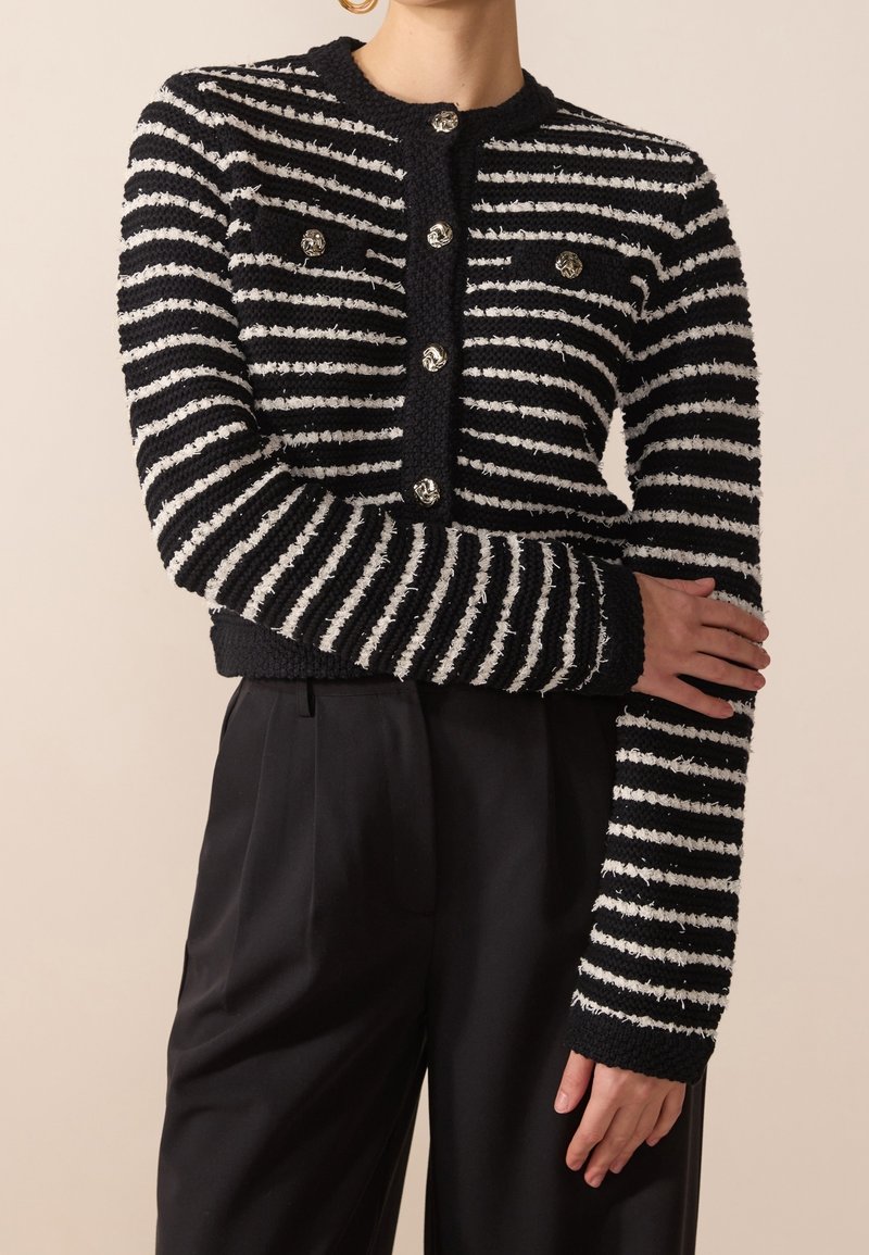 Randig svart och vit cardigan med strukturerad stickning, rundade knappar, långa ärmar och kort längd, tillsammans med svarta byxor.