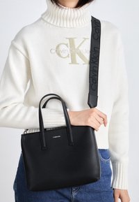 Schwarze Lederhandtasche mit zwei kurzen Griffen, einem abnehmbaren Riemen, flacher Form und silbernem Marken-Detail auf der Vorderseite.