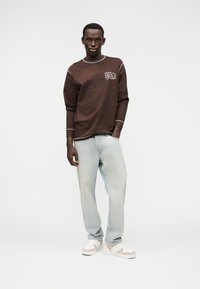 Καφέ πουκάμισο με μακριά μανίκια και λευκό γραφικό κείμενο, ανοιχτό μπλε φαρδιά τζιν και λευκά αθλητικά παπούτσια με μπεζ λεπτομέρειες, στέκεται μπροστά σε απλό φόντο.