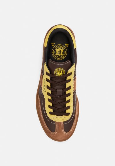 Pruun vabaajajalats kollaste ja beežide aktsentidega, tekstuurse nahaga, ümarate paeltega ja keelel olev ringikujuline logo. Sile pind tikanditega.