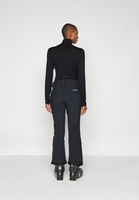 Sort langærmet top og sorte skibukser med lynlåse ved anklerne, parret med sorte skistøvler. Glat tekstur og ergonomisk pasform.