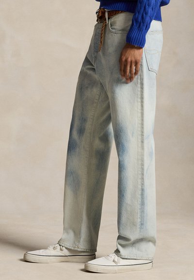 Personne portant un jean bleu clair à jambes larges avec une ceinture tressée marron, un pull bleu et des baskets blanches, debout sur un fond neutre.