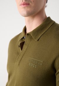 Olivno zelena polo majica z rebrastim ovratnikom, dvema gumboma in embosiranim belim logotipom "Porsche" in "BOSS" na prsih.