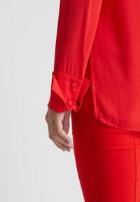 Rote Bluse mit langen Ärmeln, mit geknöpften Manschetten und einem Seitenschlitz. Kombiniert mit roten, eng anliegenden Hosen, die eine glatte Stoffoberfläche zeigen.