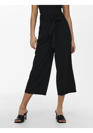 Woman wearing black wide-leg cropped pants with a belt tied at the waist and black open-toe heeled mules, standing on a white background.