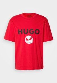T-shirt rouge à manches courtes avec texte noir "HUGO" et graphisme blanc stylisé d'un visage de crâne centré en dessous du texte.