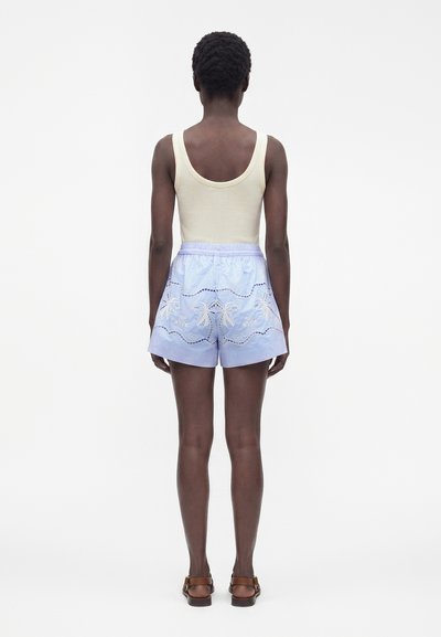 Personne debout, de dos, portant un débardeur crème, un short bleu clair brodé avec des motifs de palmiers et des sandales marron.